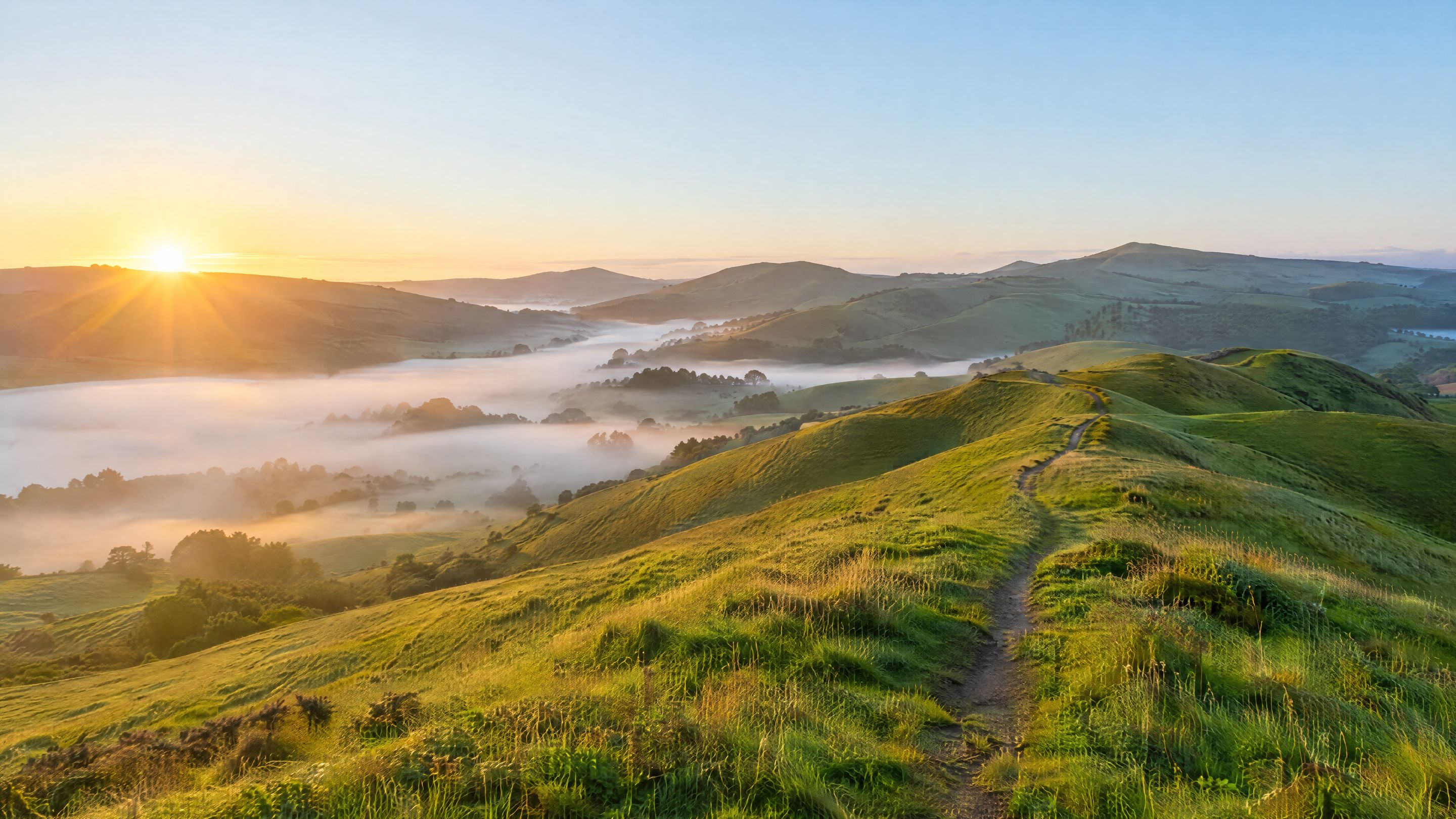 A footpath winding over green hills at sunrise, mist lying in the valleys